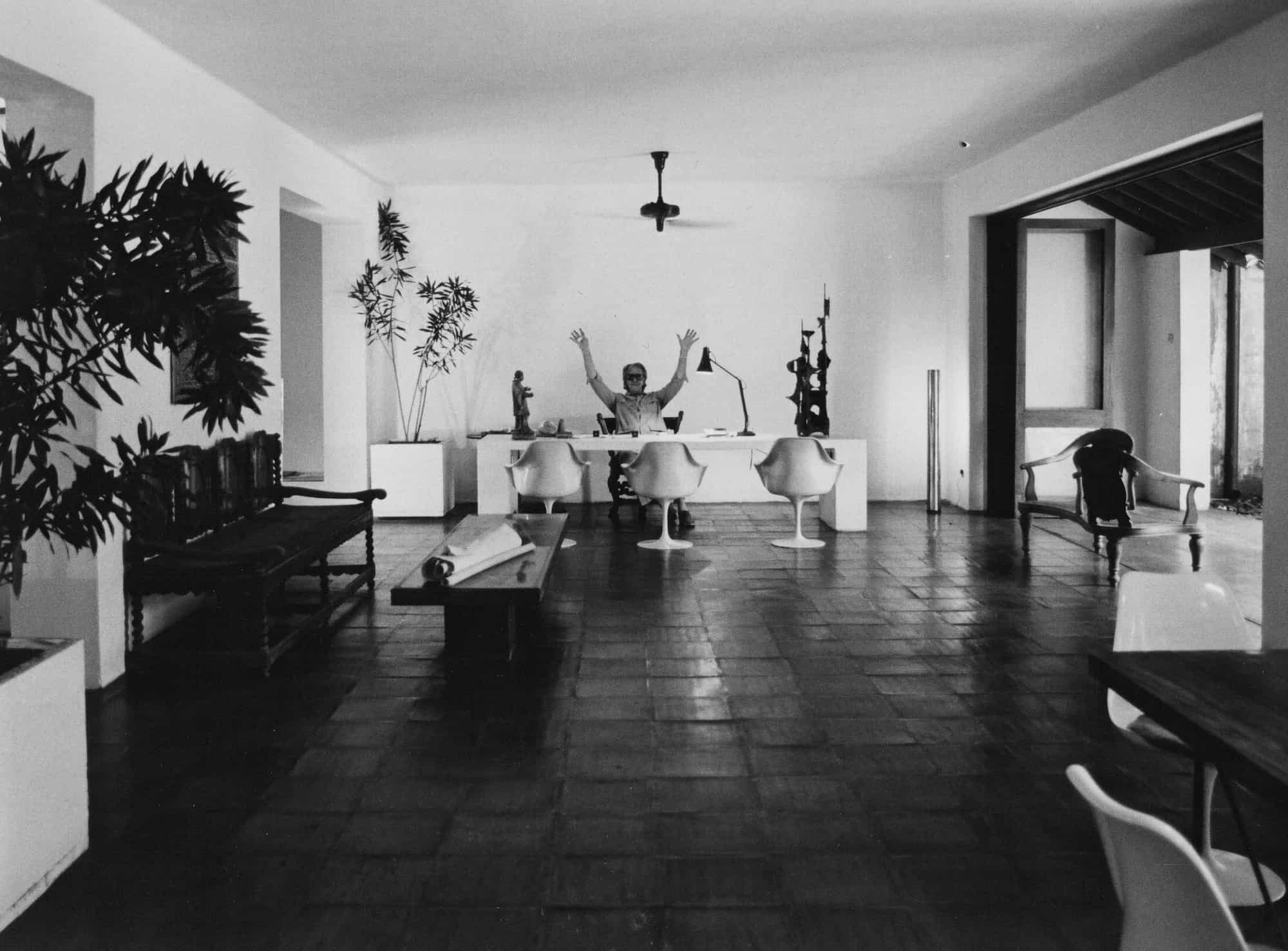 Geoffrey Bawa in his Studio, 1985, with his arms raised behind his desk at one end of the white walled open plan room