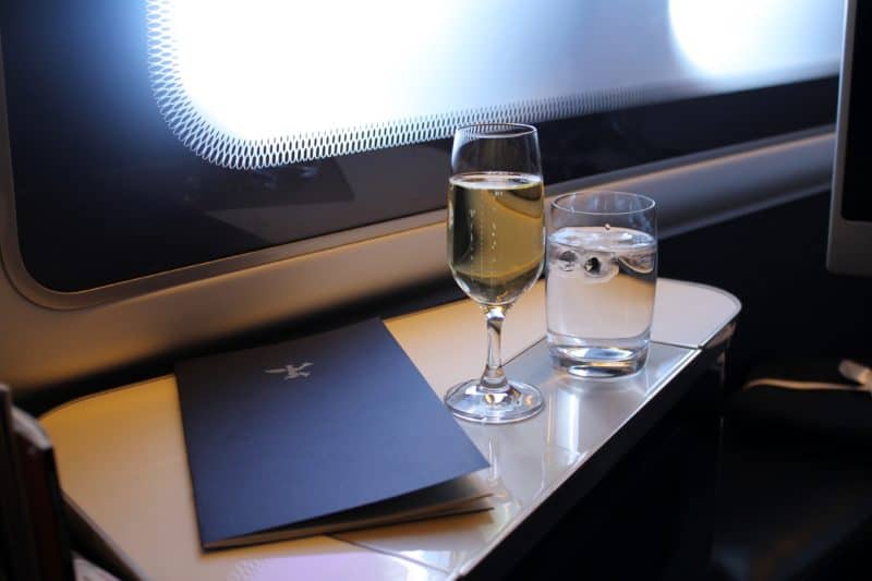 Champagne, a dark blue menu and a glass of water in a business class seat on a table next to the window