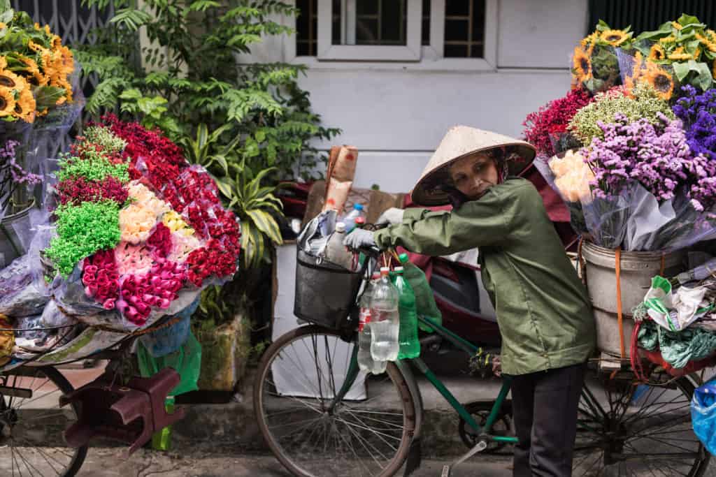 Flower hawker with conical hat pushing bike with flowers on the back
