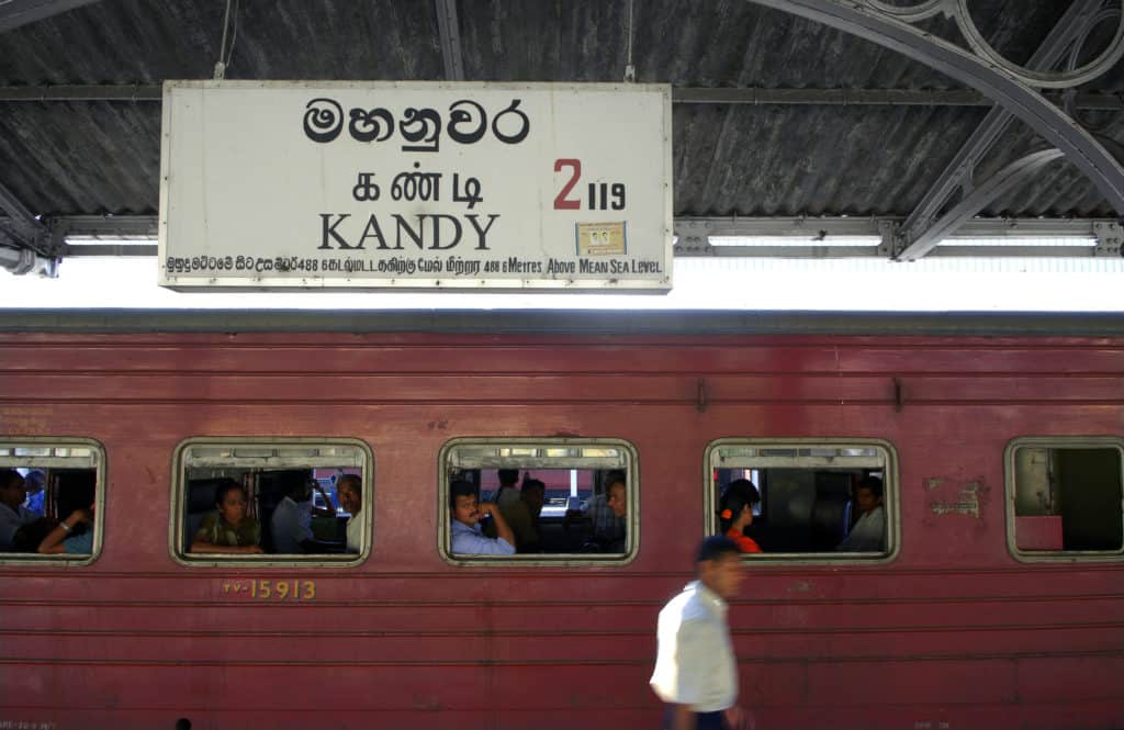 A sign at Kandy Train station with a train behind it and gentleman walking past on the platform