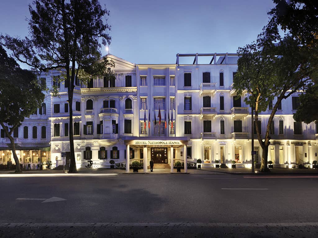 White exterior of the Sofitel Legend Metropole in Hanoi from across the road with a couple of trees in front