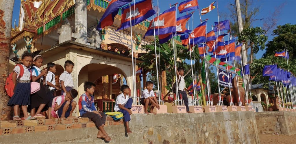 School children on steps underneath flag poles with the cambodian flag on top