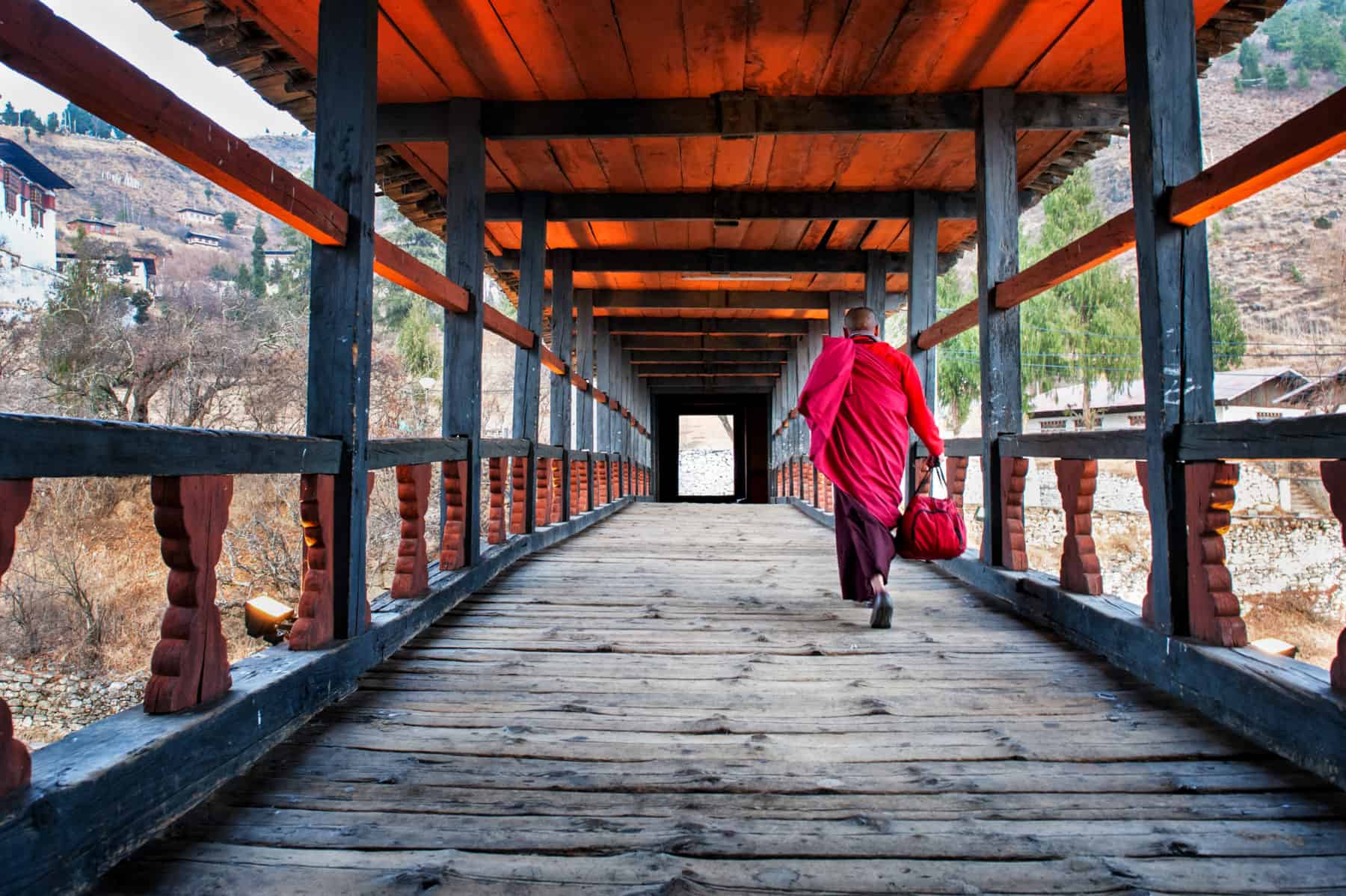 A monks dressed in red traditional costume walks across a bridge in bhutan