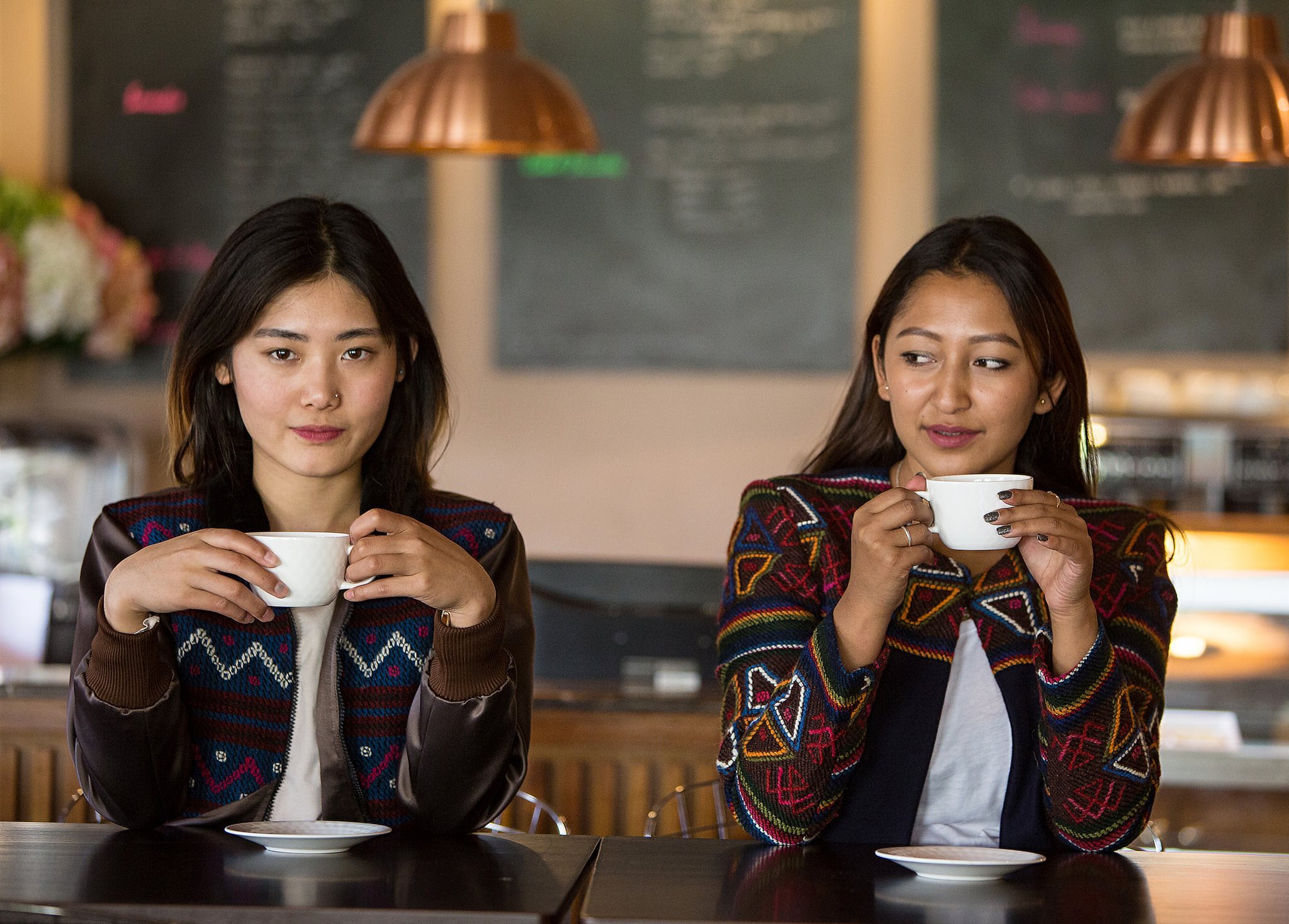 Two young Bhutanese woman sat next each other in a cafe, drinking a hot drink