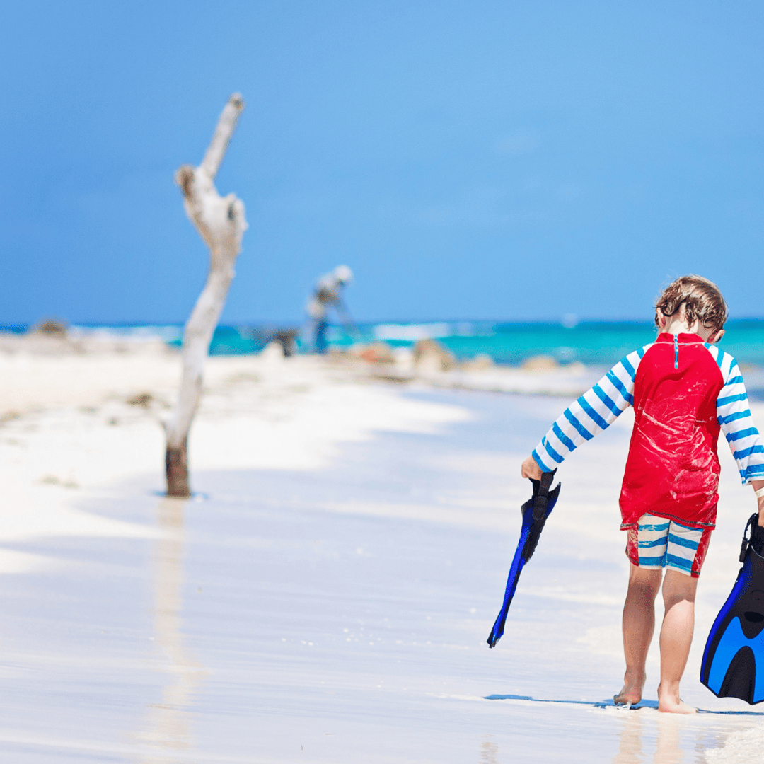 Little boy walking along a beach in the Maldives holding diving flippers.