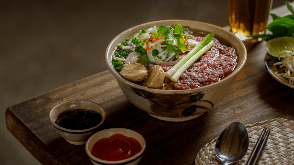 Traditional bowl of Pho noodles on a wooden table with cutlery.