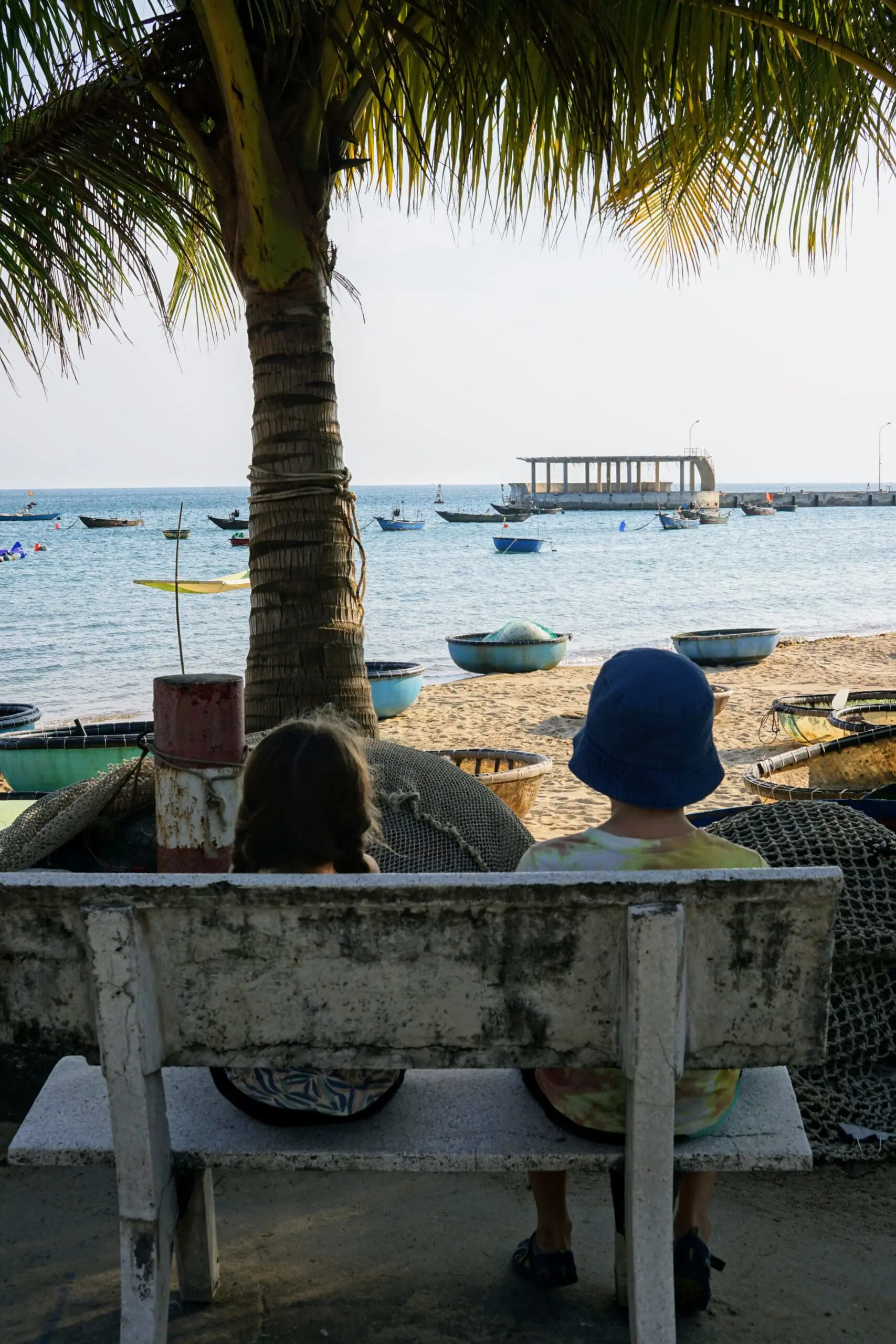 Two young kids sitting on a bench facing the sea on a beach in Hoi An